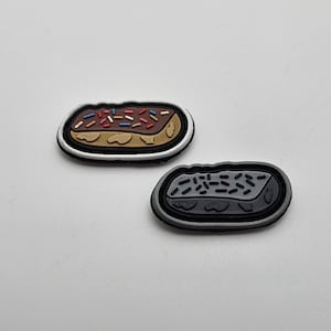 May include: Two iron-on patches shaped like eclairs. One is white with a brown and white eclair with red, blue, and white sprinkles. The other is gray with a gray eclair with black sprinkles.