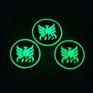 Puede incluir: Tres parches redondos, negros y que brillan en la oscuridad con un diseño de fénix blanco. El fénix se representa con las alas extendidas y un diseño de llama debajo.