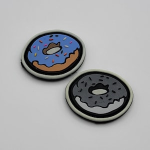 Könnte beinhalten: Zwei Donut-förmige Patches, einer blau mit rosa und weißen Streuseln und einer grau mit schwarzen Streuseln.