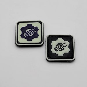 Puede incluir: Dos parches de forma cuadrada con bordes blancos. Los parches presentan un engranaje blanco con el texto "EDC" en morado sobre un fondo gris claro. El otro parche tiene un engranaje negro con el texto "EDC" en blanco sobre un fondo negro.