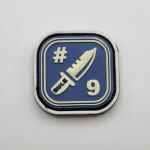 Könnte beinhalten: Ein blauer und weißer Patch mit einem weißen Messer und dem Text "RULE #9" darauf.
