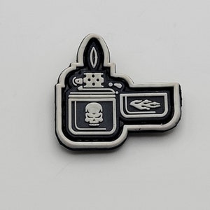 Puede incluir: Un parche blanco y negro con una ilustración detallada de un encendedor con un diseño de calavera y una llama. El encendedor está delineado en blanco y la calavera y la llama son negras.