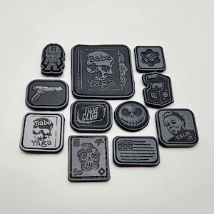 Op de afbeelding: Een verzameling van 11 zwart-witte strijklabels met verschillende ontwerpen, waaronder een schedel, een pistool, een videogame, een personage uit de film "Fight Club", een personage uit de film "Nightmare Before Christmas", een personage uit de film "Halloween", en de tekst "Baba Yaga" en "Killin' It".