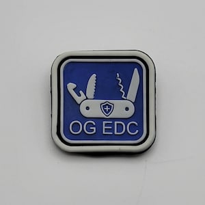 Puede incluir: Un parche azul y blanco con un contorno blanco. El parche presenta una silueta blanca de una multiherramienta con el texto "OG EDC" en blanco.