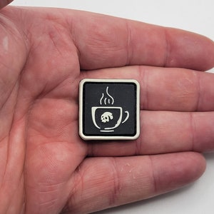 K&ouml;nnte beinhalten: Ein schwarzer quadratischer Aufn&auml;her mit wei&szlig;em Rand, der ein wei&szlig;es Kaffeetassen-Design mit einem Totenkopf im Inneren zeigt. Dampf steigt aus der Tasse auf. Der Aufn&auml;her wird in einer Hand gehalten.