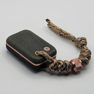 May include: A small, dark green, rectangular speaker with a copper-colored trim and a copper-colored button on the top. The speaker is attached to a brown and white braided rope with a copper-colored bead.
