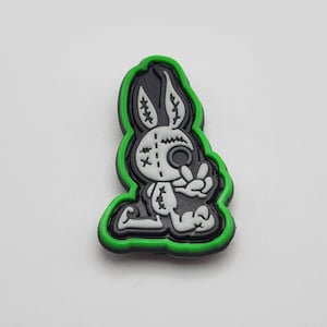 May include: A black and white cartoon bunny rabbit with stitched seams and a single eye is outlined in green. The bunny is kneeling with its hands clasped together.
