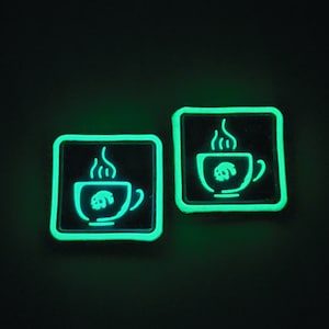 K&ouml;nnte beinhalten: Zwei quadratische, gr&uuml;n leuchtende Aufn&auml;her auf schwarzem Hintergrund. Jeder Aufn&auml;her zeigt einen wei&szlig;en Umriss einer Kaffeetasse mit Dampf und einem Totenkopf-Design im Inneren. Diese Aufn&auml;her sind wahrscheinlich f&uuml;r Kleidung oder Accessoires.