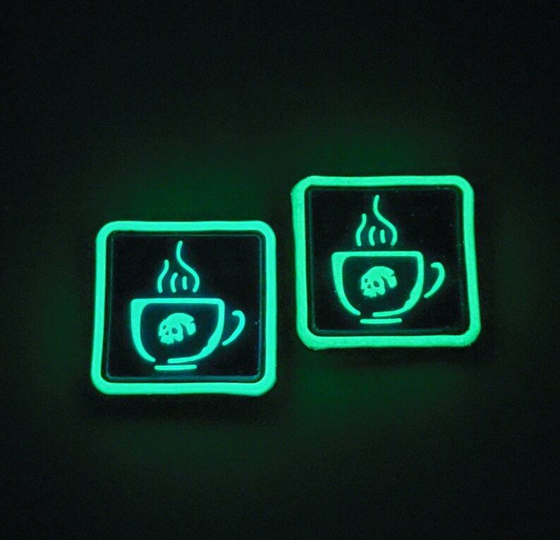 K&ouml;nnte beinhalten: Zwei quadratische, gr&uuml;n leuchtende Aufn&auml;her auf schwarzem Hintergrund. Jeder Aufn&auml;her zeigt einen wei&szlig;en Umriss einer Kaffeetasse mit Dampf und einem Totenkopf-Design im Inneren. Diese Aufn&auml;her sind wahrscheinlich f&uuml;r Kleidung oder Accessoires.
