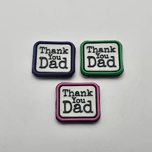Può includere: Tre toppe quadrate con sfondo bianco e testo nero che recita "Thank You Dad". Le toppe hanno bordi di colori diversi: blu, verde e rosa.