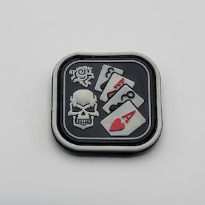 Puede incluir: Un parche gris y blanco con un diseño de calavera y huesos cruzados y cartas de juego. Las cartas son un as de corazones, una reina de diamantes, un jack de tréboles y un rey de picas. El parche tiene un borde negro y un contorno blanco.