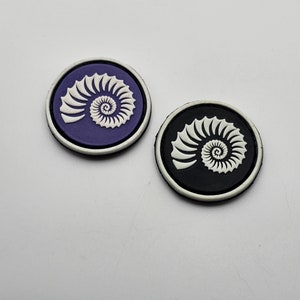 Peut inclure: Deux patchs ronds en tissu avec un motif de coquille de nautile blanche. Un patch est violet et l'autre est noir.