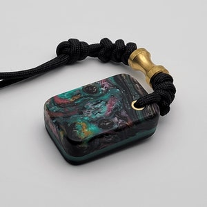 May include: A black cord with a brass bead and a rectangular pendant. The pendant is made of a green, black, and pink resin with a swirling pattern.