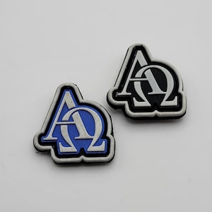 Op de afbeelding: Twee schoenbedels met de Griekse letters Alpha en Omega. Eén bedel heeft een blauwe achtergrond met witte letters en een zwarte rand. De andere heeft een zwarte achtergrond met witte letters en een zwarte rand.