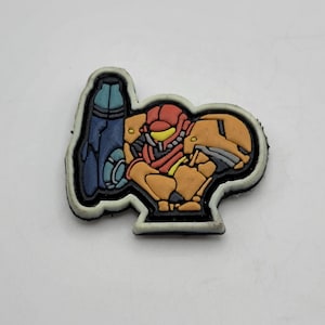 May include: A black and white patch with a cartoon image of a red, orange, and yellow character from the video game Metroid. The character is holding a blue and white weapon.