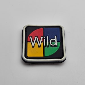 May include: A white, black and red embroidered patch with the word "Wild" in white letters on a black background. The patch is surrounded by a white border.