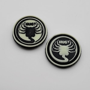 May include: Two black and white round buttons with a glow-in-the-dark alien creature design. The alien has a large head and long, thin legs. The text "HUG?" is printed above the alien on each button.