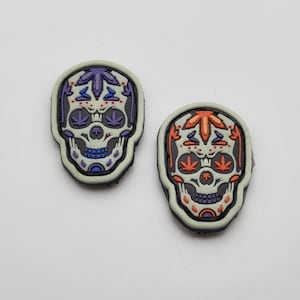 Puede incluir: Dos parches de calavera de azúcar coloridos con fondo blanco y contornos negros. Las calaveras tienen un diseño detallado con acentos naranja, morado y azul. Los ojos están delineados en azul y tienen un diseño de hoja de marihuana verde en el interior.
