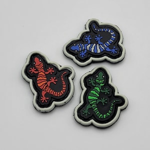 May include: Three iron-on patches featuring different colored lizards. The first patch is blue and white, the second is orange and red, and the third is green. All three patches are outlined in black.