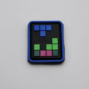 Puede incluir: Un parche azul y negro con un diseño tipo Tetris. El diseño presenta un bloque en forma de T y una fila de bloques en verde, rosa y negro.