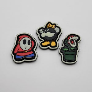 Puede incluir: Tres parches bordados para planchar con personajes de la serie de videojuegos Super Mario Bros. Los parches son rojo, negro y verde. El parche rojo es de un personaje con una máscara, el parche negro es de un personaje con una corona y el parche verde es de una planta con una boca.