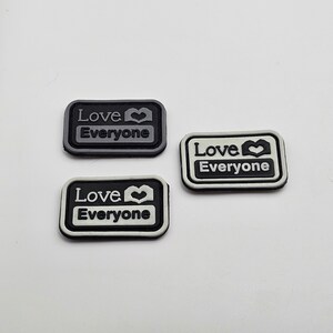 May include: Three rectangular patches with the words "Love Everyone" and a heart symbol. The patches are in black, white, and gray, with a black border. The text and heart are in white.