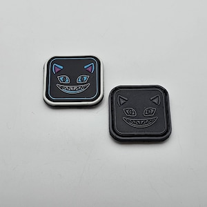 May include: Two black and white square patches with a cartoon cat face with a mischievous grin. The cat has blue eyes and pink ears. The patches are made of a soft, textured material.
