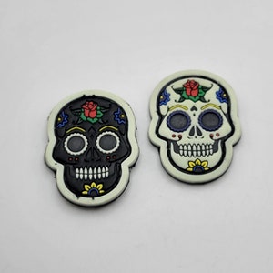 May include: Two sugar skull patches, one black and one white, with colorful floral designs. The skulls have black eyes and a white smile.