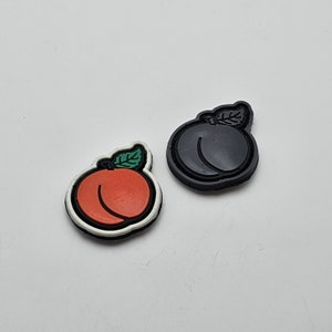 Könnte beinhalten: Zwei gummiförmige Pflaster in Form von Pfirsichen, eines schwarz und eines weiß mit einem roten Pfirsich-Design.
