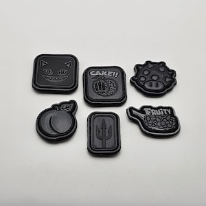 Op de afbeelding: Zes zwarte strijkpatches met verschillende ontwerpen. De patches bevatten een perzik, een drietand, een stekelig wezen, een kattengezicht, een perzik met de tekst "CAKE!" en een tank met de tekst "FRUITY".