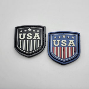 May include: Two embroidered patches in the shape of a shield with the word "USA" in white letters and a row of stars above the word. The patch on the left is black and white, and the patch on the right is blue with red and white stripes.