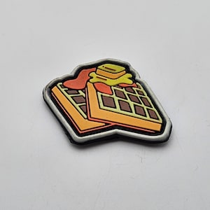 May include: A white, brown, and yellow waffle patch with a white border. The waffle is topped with a yellow pat of butter and a red syrup drizzle.