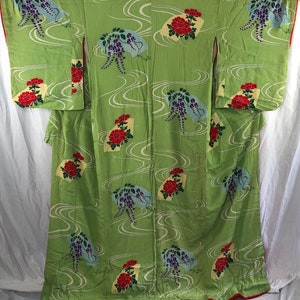 May include: A light green kimono with a repeating pattern of red flowers, purple wisteria, and fan-shaped designs. The kimono has a red trim and white wavy lines throughout the fabric.