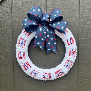 May include: A patriotic wreath with a white, fluffy base and a navy blue bow adorned with red and white stars. The wreath features red, white, and blue ribbon accents, creating a festive and decorative piece.