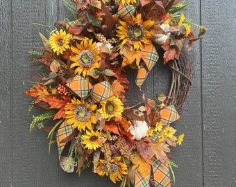 May include: A fall-themed wreath with sunflowers, leaves, and plaid ribbon. The wreath is made on a grapevine base and is decorated with faux sunflowers, leaves, and berries. The wreath is tied with a plaid ribbon.