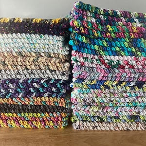 May include: A stack of colorful crocheted dishcloths, some with a speckled pattern, others with a more solid color. The dishcloths are made of cotton yarn and are perfect for cleaning up spills and messes in the kitchen.