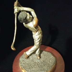May include: A silver-toned figurine of a golfer in mid-swing, standing on a round, brown wooden base. The base is inscribed with the name "C.A. Parcell".