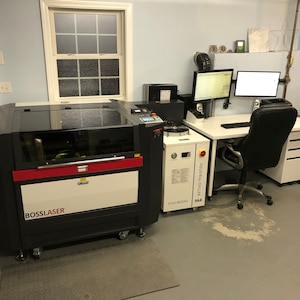 May include: A black and red laser cutter with a white label that says "BOSSLASER" is in a workshop setting. The laser cutter is next to a white industrial chiller labeled "INDUSTRIAL CHILLER CW-5000 K.A.A.". A white desk with a black office chair is in the background.