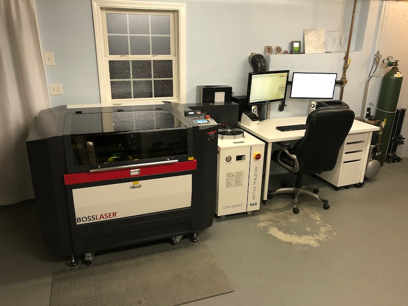 May include: A black and red laser cutter with a white label that says "BOSSLASER" is in a workshop setting. The laser cutter is next to a white industrial chiller labeled "INDUSTRIAL CHILLER CW-5000 K.A.A.". A white desk with a black office chair is in the background.