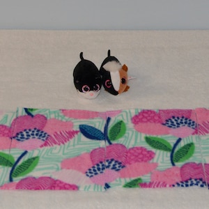 May include: A pink and green floral fabric with a repeating pattern of flowers and leaves. The fabric is laid out flat on a white surface. Two small plush toys are in the background.