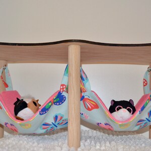 May include: Two hanging fabric hammocks with butterfly patterns, suspended from a wooden stand. One hammock has a brown and white stuffed animal inside, and the other has a black and white stuffed animal inside.