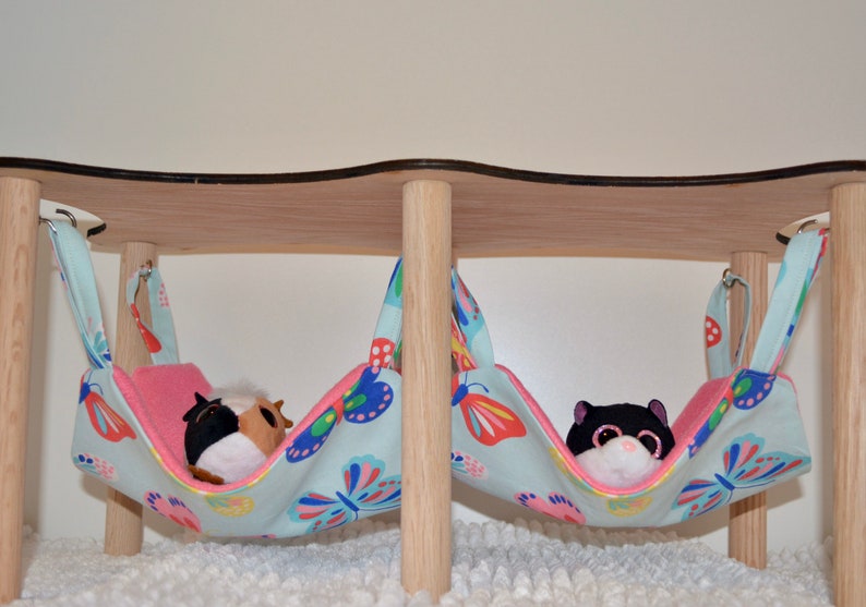 May include: Two hanging fabric hammocks with butterfly patterns, suspended from a wooden stand. One hammock has a brown and white stuffed animal inside, and the other has a black and white stuffed animal inside.