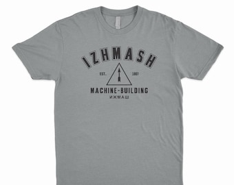 May include: A gray t-shirt with a black graphic design. The graphic features the text "IZHMASH" in a vintage collegiate style font, with the text "EST. 1807" below it. A black triangle with an arrow pointing upwards is in the center of the graphic. The text "MACHINE-BUILDING" is below the triangle, and the text "ИЗМАШ" is below that.