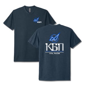 May include: A dark blue short-sleeve t-shirt featuring a small logo on the front left chest and a large graphic on the back. The graphic includes a blue and white emblem, the Cyrillic text KBP, and the text KONSTRUKTORSKOYE BYURO PRIBOROSTROYENIYA TULA, ROSSII.