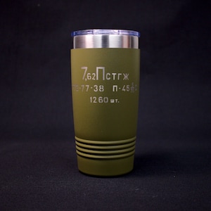 May include: Olive green travel tumbler with a stainless steel rim and a clear lid. The tumbler features engraved text in a military style font. The text includes the numbers "7,62" and other Cyrillic characters. The bottom of the tumbler has three horizontal grooves.