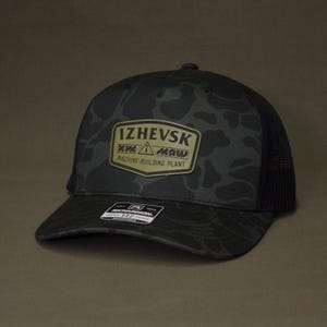 May include: Black camouflage pattern baseball cap with a mesh back. The cap features a patch with the text "Izhevsk Machine-Building Plant" in gold and white. The brim is curved and the cap has a button on top.