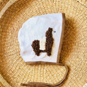May include: A white baby bonnet with a brown and white llama applique. The bonnet has a brown trim and ties.