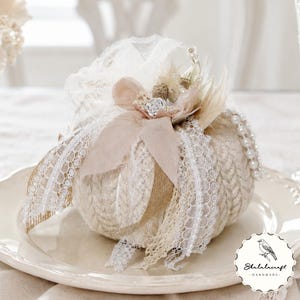 May include: A cream-colored fabric pumpkin decoration featuring a textured knit pattern, adorned with white lace ribbons, a beige fabric flower, small faux pearls, and dried botanical elements. A circular logo with a bird and the text Shalalacraft Handmade.