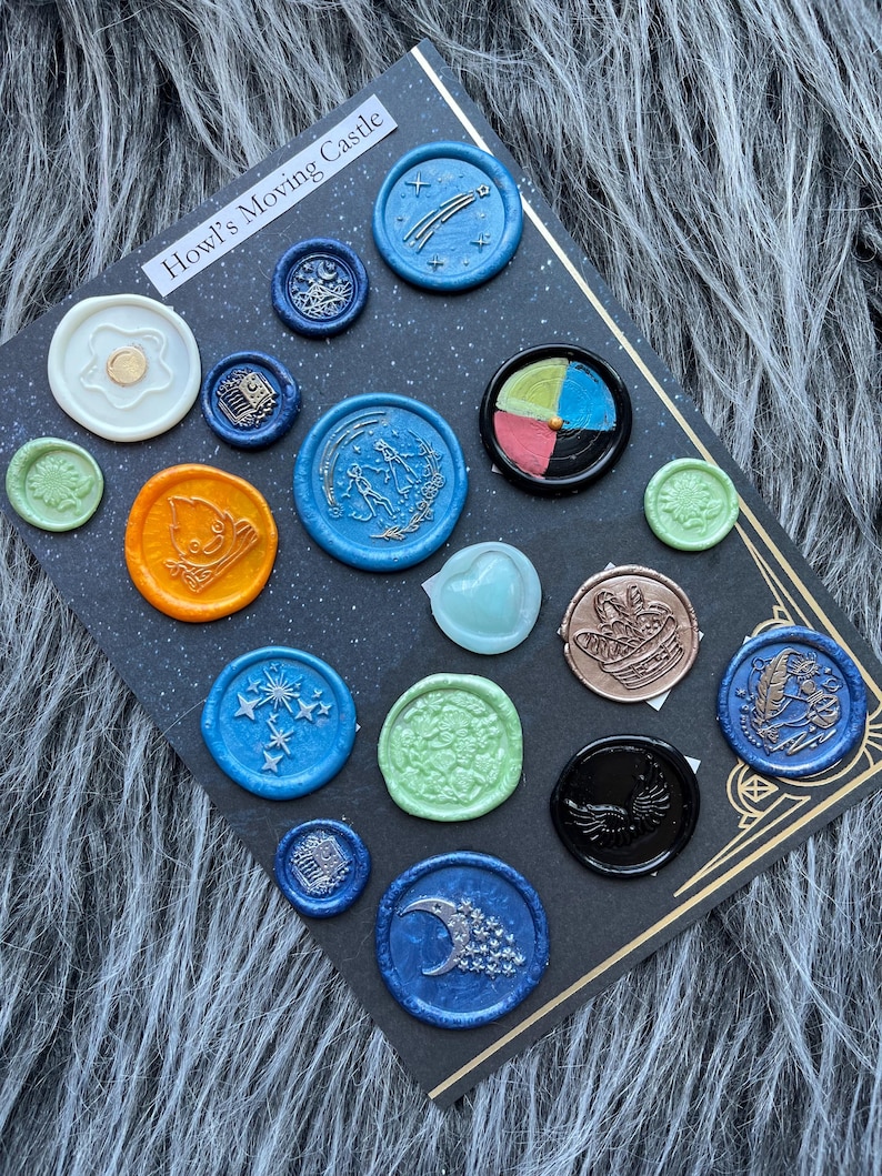 May include: A collection of colorful wax seals with various designs, including stars, moons, and abstract shapes. The seals are arranged on a black card with the text "Howl's Moving Castle". The seals are in shades of blue, orange, green, and black.