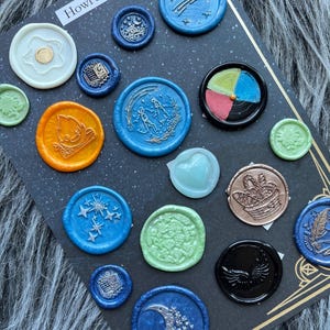 May include: A collection of colorful wax seals with various designs, including stars, moons, and floral patterns. The seals are arranged on a dark background, with the text "Howl's Moving Castle" visible at the top.